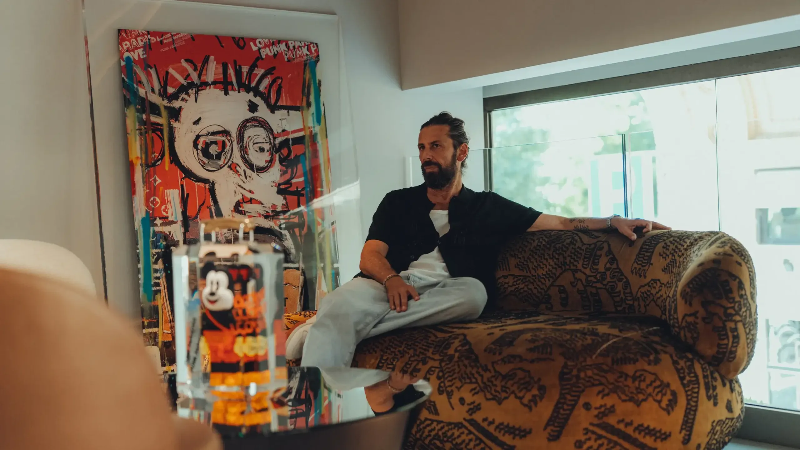Fred Allard, bearded, in a black shirt and light jeans, sitting on a patterned sofa beside a large red painting, with a Mickey Mouse figure in resin in the foreground