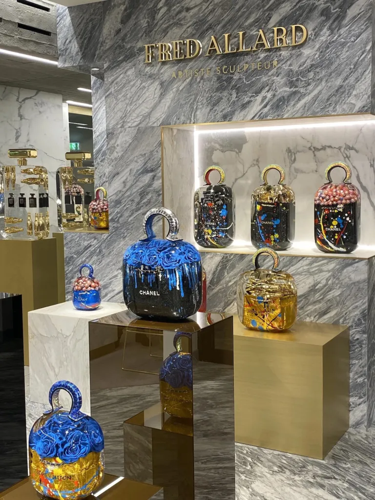 Resin sculptures of Chanel, Dior and Louis Vuitton bags on marble and brass plinths, below a gold sign reading Fred Allard, Artiste Sculpteur