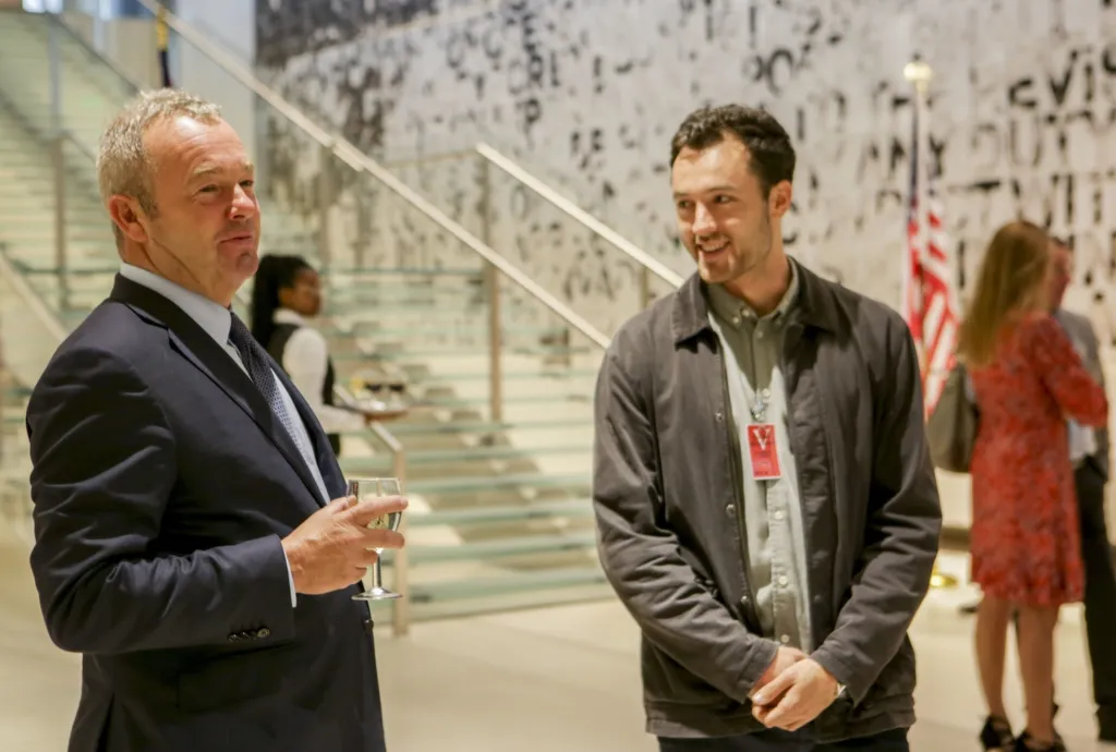 David Yarrow in a dark suit holding a glass of white wine, talking with a smiling young man at a reception by a glass staircase