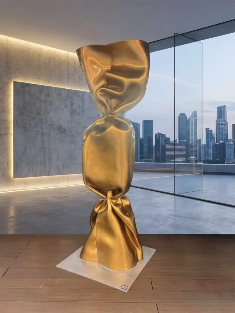 A tall gold Wrapping Bonbon sculpture on a square base in a room with concrete walls and a city skyline beyond the windows